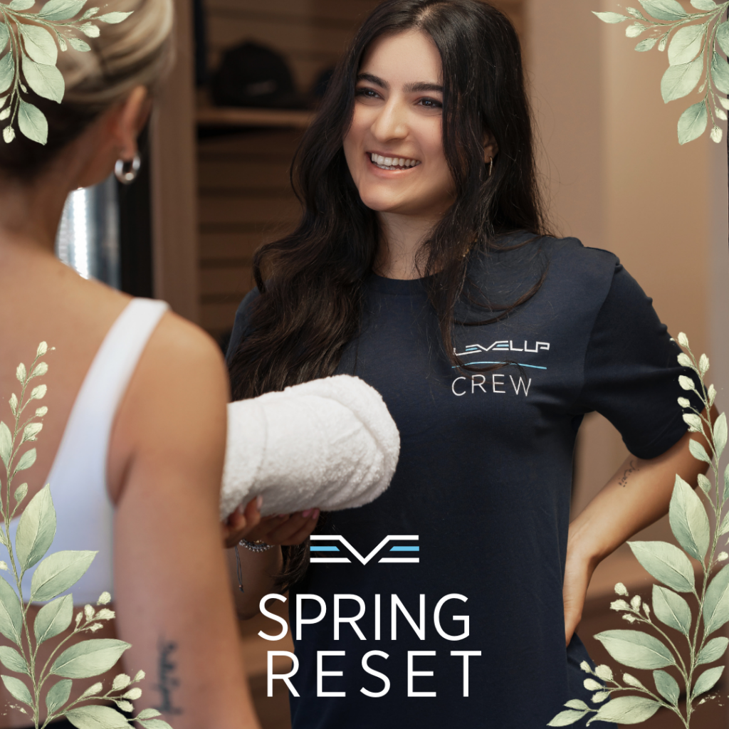 Level Up crew member smiling while handing a towel to a gym member, representing a welcoming fitness environment, with soft botanical spring elements around the image for the Spring Reset campaign.